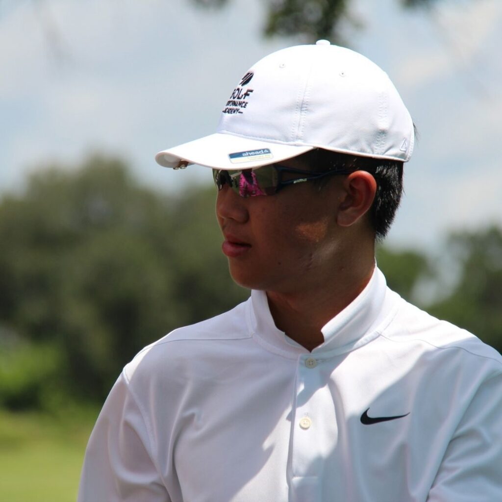 Close-up profile view of a young male junior golfer in sunglasses and a white polo shirt looking focused on the golf course.