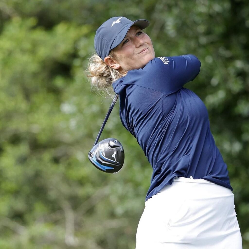 Female golfer wearing a blue shirt and white skirt powerfully hitting a driver off the tee, captured mid-swing.