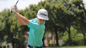 Young male junior golfer wearing a white Titleist hat and a striped turquoise shirt mid-swing with a driver on a sunny course.