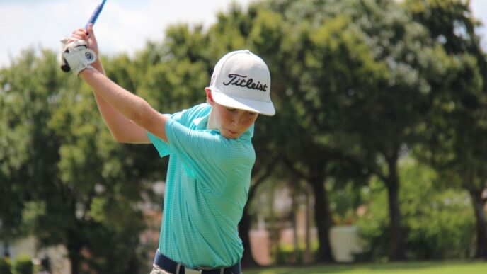 Young male junior golfer wearing a white Titleist hat and a striped turquoise shirt mid-swing with a driver on a sunny course.